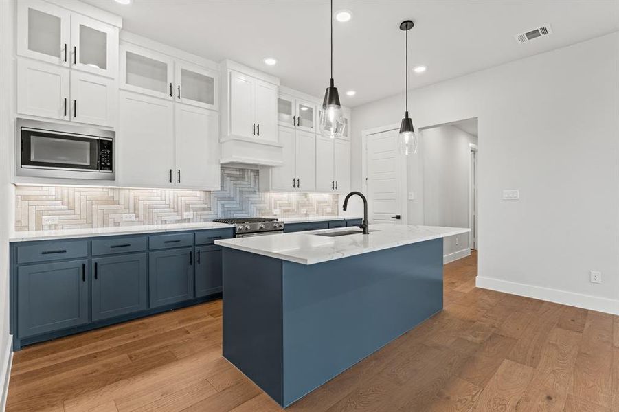 Kitchen with tasteful backsplash, built in microwave, sink, white cabinetry, and an island with sink