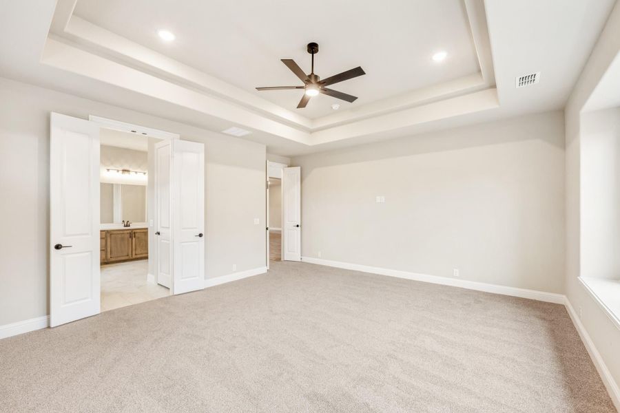 Spacious master bedroom with tray ceiling, ceiling fan, carpet flooring, and ensuite bathroom access. Spacious master bedroom with tray ceiling, ceiling fan, carpet flooring, and ensuite bathroom access.