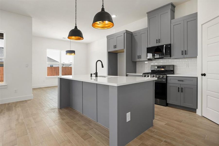 Kitchen featuring appliances with stainless steel finishes, a center island with sink, hanging light fixtures, gray cabinets, and decorative backsplash