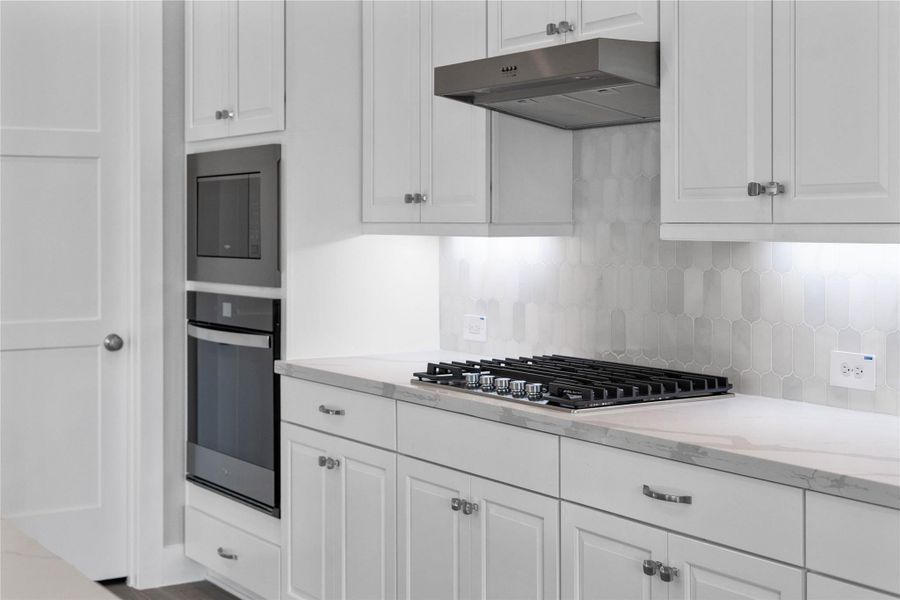 Top-of-the-line stainless steel appliances that offer both professional function and modern style. The focal point is a professional-grade gas cooktop with multiple burners and robust grates, perfectly complemented by an elegant stainless steel range hood. A built-in microwave conveniently located above the coordinating built-in oven, creating a highly efficient and visually appealing cooking center that streamlines meal preparation.