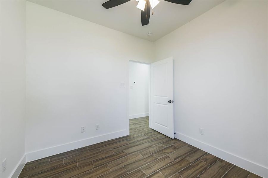 This is a bright, empty room with clean white walls and modern wood-look tile flooring. There's a white door on the right, and the room features a contemporary ceiling fan with lighting.