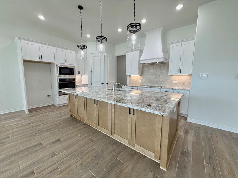 Spacious kitchen featuring a large island with a stone countertop, wood-finish flooring, white upper and lower cabinetry, stainless steel built-in appliances, and recessed lighting