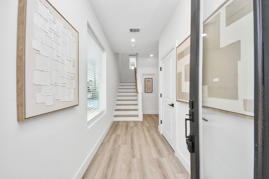 This photo showcases a bright and welcoming entryway with light wood flooring and modern art on the walls. It features a staircase leading to the upper level and a large window providing natural light. The overall design is clean and contemporary.