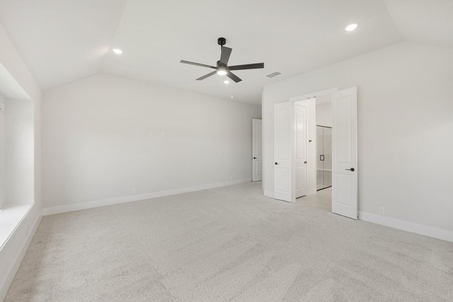 Empty bedroom with vaulted ceiling, ceiling fan, carpet flooring, and open doors to ensuite bathroom