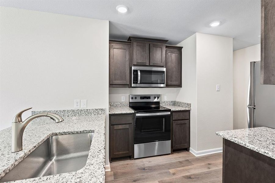 Kitchen featuring stainless steel appliances, dark brown cabinetry, light wood finished floors, and light stone counters