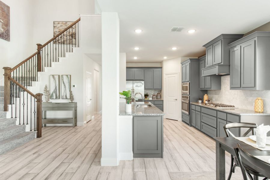 Open-concept kitchen with gray cabinets and island, adjacent to staircase with wood railings and light hardwood floors.