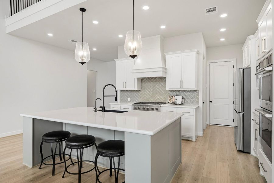 The island kitchen makes a striking statement with sleek Quartz countertops, a deep granite sink and decorative pendant lighting above the breakfast bar. A chevron-pattern subway tile back splash adds custom visual interest, while recessed lighting enhances the clean, contemporary design and highlights the kitchen’s refined finishes.