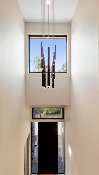 The photo showcases a modern entryway with high ceilings and a large window above the door, allowing natural light to fill the space. A stylish, contemporary chandelier adds elegance to the minimalist design.