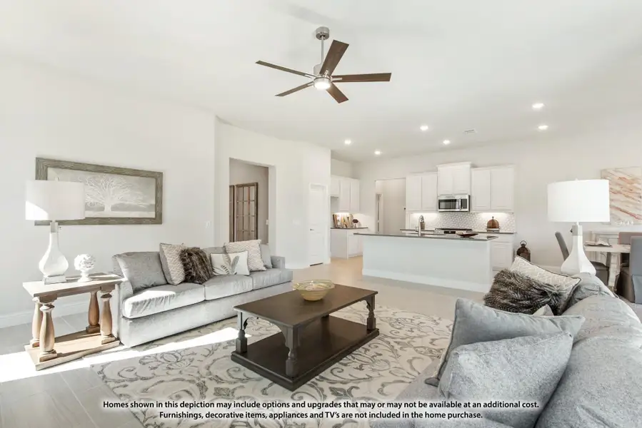 Open-concept living room with gray sofas and ceiling fan, open to white kitchen with island in background. Open-concept living room with gray sofas and ceiling fan, open to white kitchen with island in background.