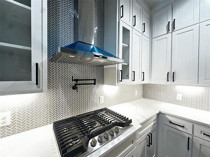 Kitchen with glass insert cabinets, stainless steel gas stovetop, wall chimney exhaust hood, light stone countertops, and white cabinets Kitchen with glass insert cabinets, stainless steel gas stovetop, wall chimney exhaust hood, light stone countertops, and white cabinets