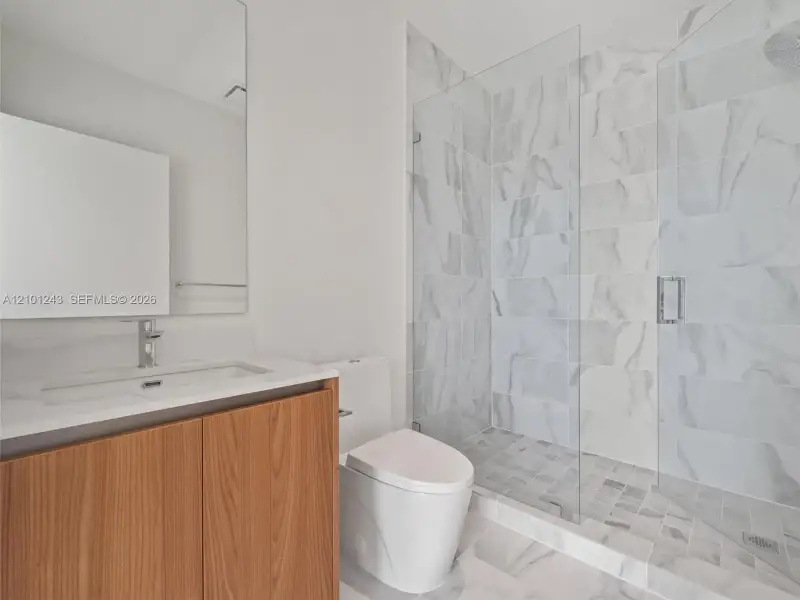 Bathroom featuring a vanity with a light-toned wood-finish cabinet and solid-surface countertop, a frameless glass-enclosed shower with large-format wall tile, and a wall-mounted mirror Bathroom featuring a vanity with a light-toned wood-finish cabinet and solid-surface countertop, a frameless glass-enclosed shower with large-format wall tile, and a wall-mounted mirror