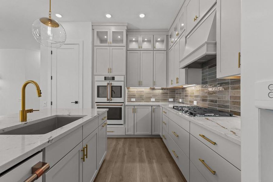 Kitchen featuring backsplash, decorative light fixtures, light stone countertops, white double oven, and custom range hood