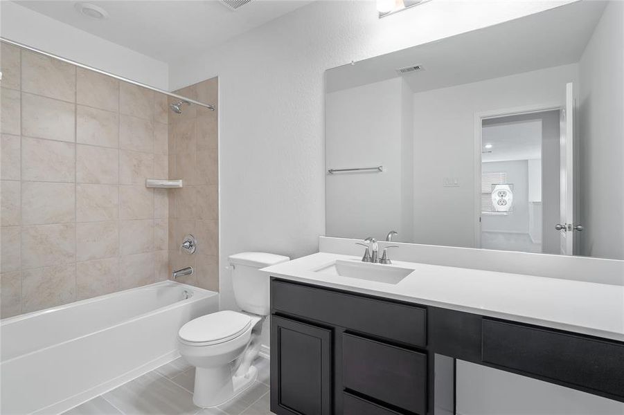 Secondary bath features tile flooring, bath/shower combo with tile surround, dark stained wood cabinets, beautiful light countertops, mirror, dark, sleek fixtures and modern finishes.