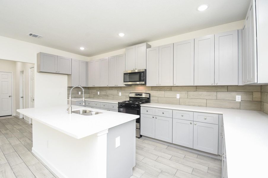 This is a spacious, modern kitchen featuring white countertops, ample light grey cabinetry, stainless steel appliances, and a central island with a sink. The room has wood-look tile flooring and is well-lit with both natural and artificial light. This is a spacious, modern kitchen featuring white countertops, ample light grey cabinetry, stainless steel appliances, and a central island with a sink. The room has wood-look tile flooring and is well-lit with both natural and artificial light.