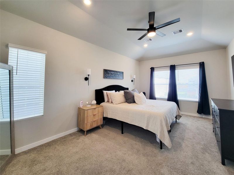 This spacious Primary bedroom features neutral walls, a cozy carpet, and a ceiling fan. It includes a large window with curtains, providing ample natural light, and is furnished with a comfortable bed, nightstand, and dresser.
