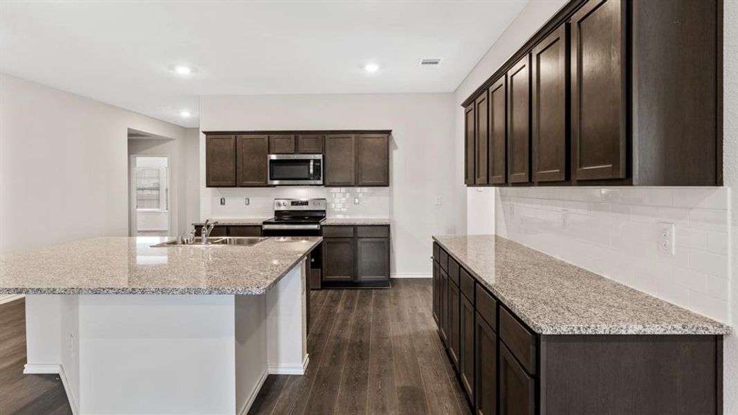 Kitchen with dark brown cabinetry, appliances with stainless steel finishes, light stone countertops, decorative backsplash, and dark wood-style flooring