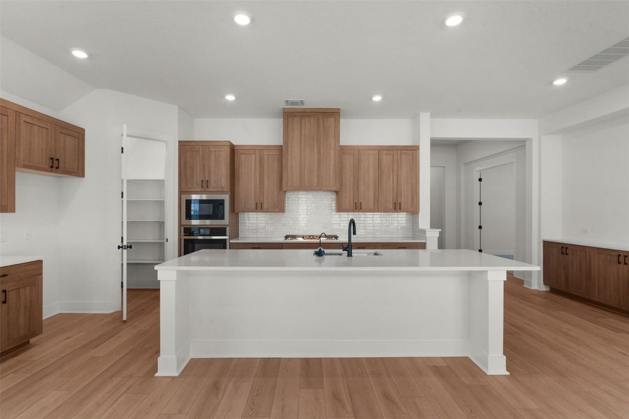 Kitchen featuring a substantial central island with an undermount sink, wood-finish flooring, recessed lighting, shaker-style cabinetry, and built-in stainless steel appliances Kitchen featuring a substantial central island with an undermount sink, wood-finish flooring, recessed lighting, shaker-style cabinetry, and built-in stainless steel appliances