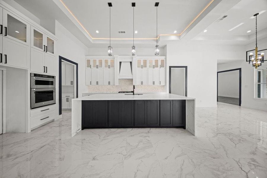 Two tone kitchen featuring glass insert cabinets, light stone countertops, a large island, a raised ceiling, and two tone cabinets