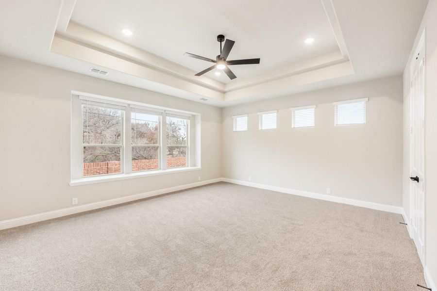 Empty bedroom with tray ceiling, ceiling fan, carpet flooring, and large windows with transom windows above Empty bedroom with tray ceiling, ceiling fan, carpet flooring, and large windows with transom windows above