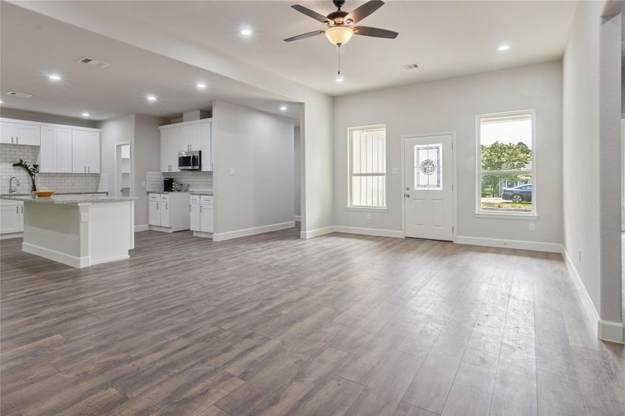 A spacious open-concept living area with modern flooring, recessed lighting, and a ceiling fan. The room connects seamlessly to a bright kitchen with white cabinetry, a large island. Natural light floods in through large windows and the glass-paneled front door.