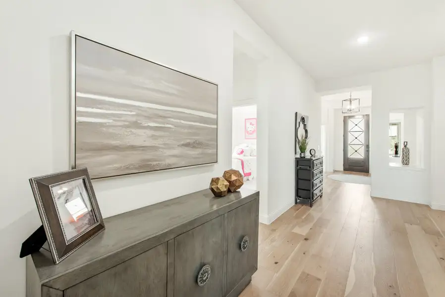 Hallway with hardwood floors, gray console table, wall art, and front door with glass panels visible in background. Hallway with hardwood floors, gray console table, wall art, and front door with glass panels visible in background.