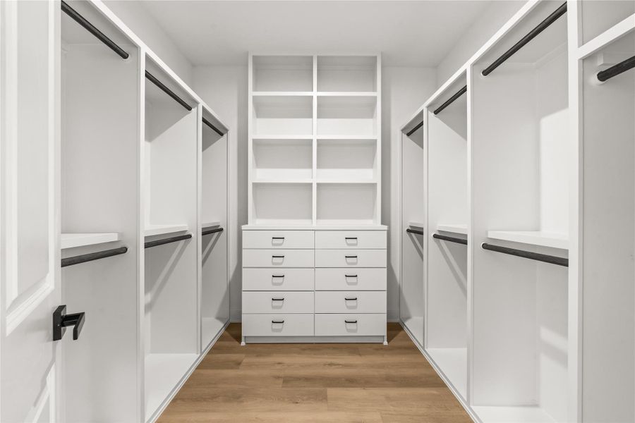 The massive primary walk-in closet features a white custom storage system and continuous light oak flooring. A central built-in dresser offers ten drawers with display shelving above, flanked by multiple wardrobe sections equipped with sturdy black hanging rods.