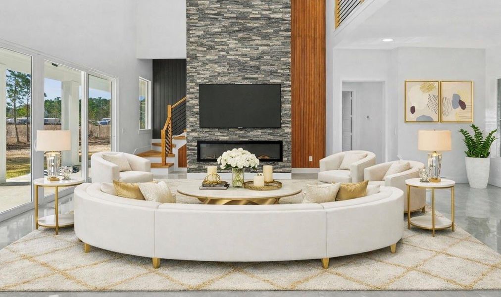 Modern living room featuring a curved white sectional sofa with gold accents centered around a round coffee table. A stone fireplace with a mounted TV serves as the focal point, complemented by elegant side tables, lamps, and neutral décor. Large windows fill the space with natural light, creating a bright and luxurious atmosphere. VIRTUALLY STAGED