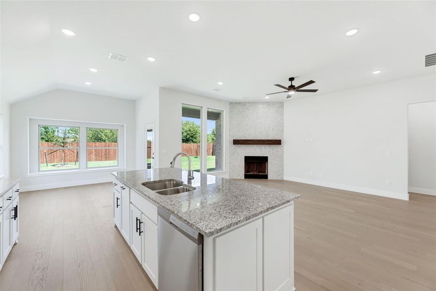 Open-concept kitchen with granite island and sink, flowing into living room with fireplace and light wood floors