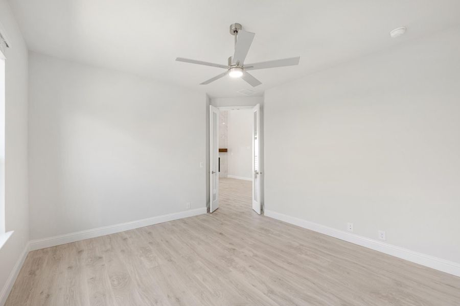 Empty bedroom with light wood floors, ceiling fan, and open double doors leading to bathroom