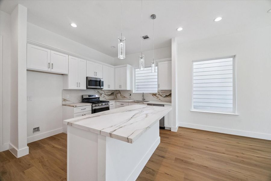 The kitchen’s oversized island becomes a natural centerpiece for gathering, dining, and meal preparation. Marble-look countertops pair beautifully with crisp white cabinetry, stainless steel appliances, and sleek modern hardware. Pendant lights add a decorative touch above the island, while recessed lighting and large windows keep the room bright. Warm wood-look flooring brings balance and warmth to the contemporary palette.