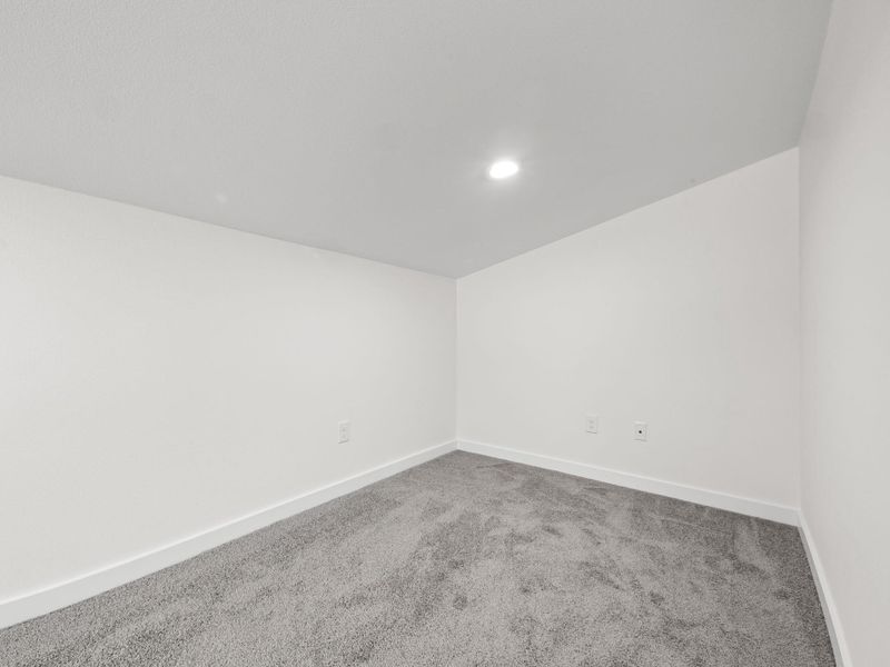 Recessed ceiling lighting illuminates a room featuring light gray carpet, white walls, and white baseboards