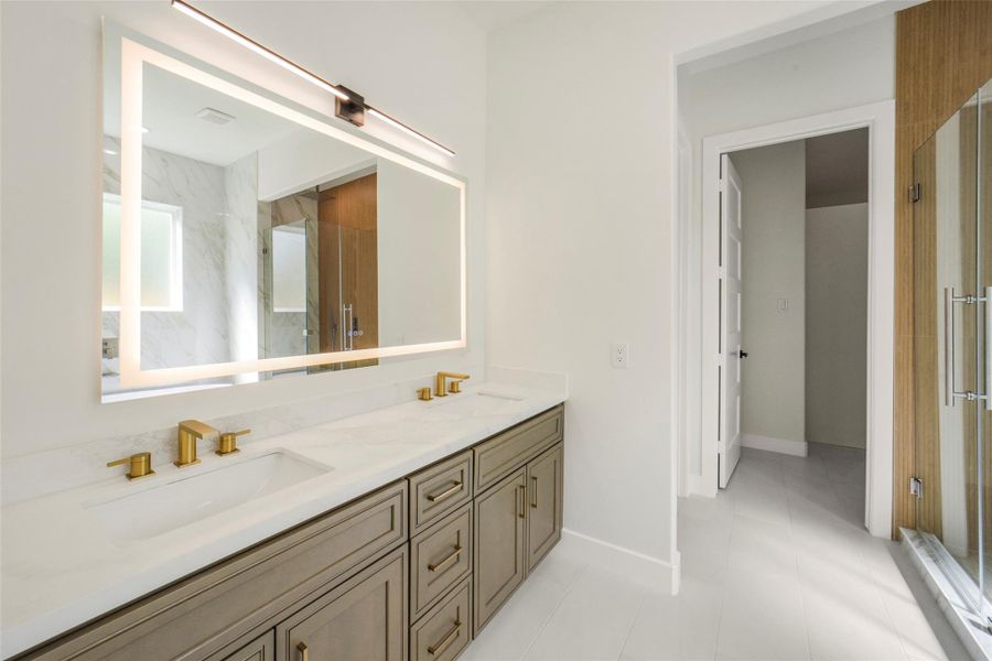 This modern bathroom features a spacious double vanity with gold fixtures, a large illuminated mirror, and a glass-enclosed shower. The neutral color palette and sleek design create a luxurious and inviting atmosphere.