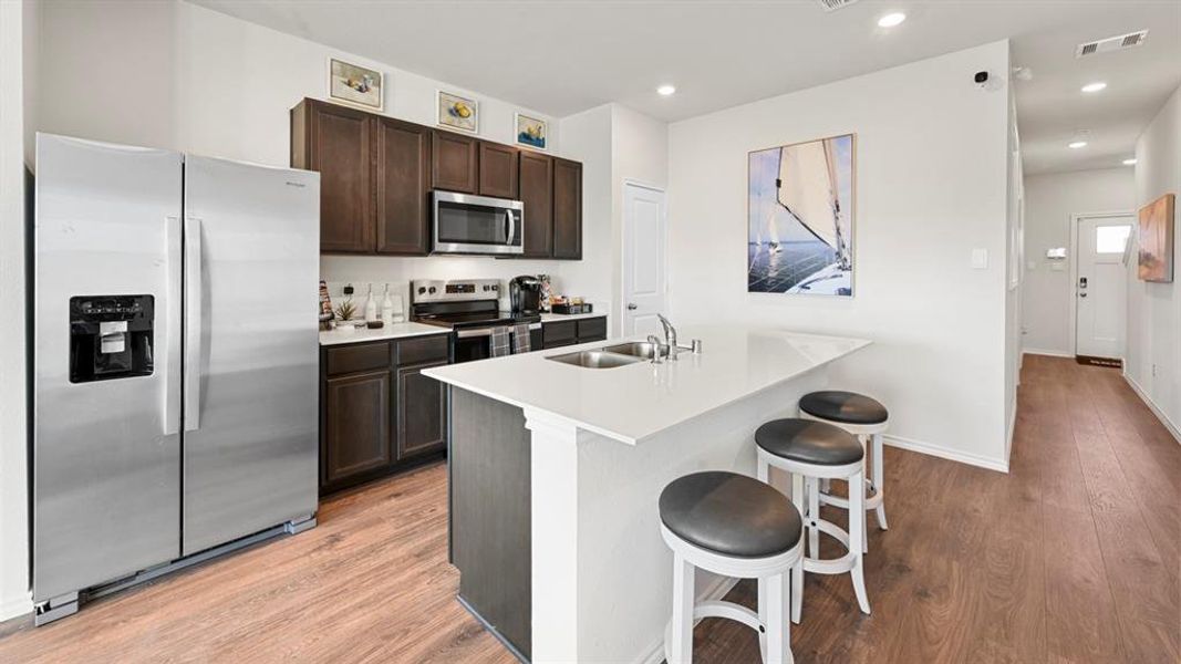 Kitchen featuring stainless steel appliances, light wood-style flooring, dark brown cabinetry, a kitchen bar, and a kitchen island with sink