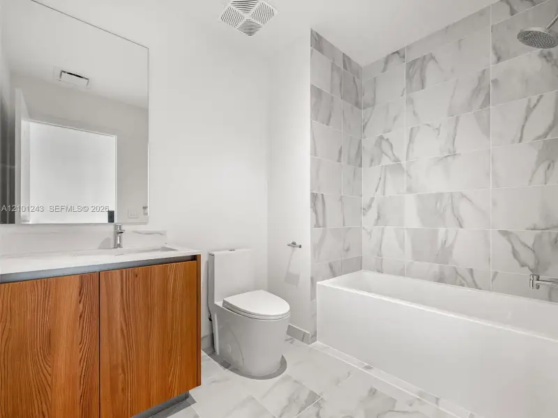 Bathroom featuring a wood-finish vanity with a rectangular sink, large frameless mirror, white plumbing fixtures, and floor-to-ceiling tile surrounding the bathtub Bathroom featuring a wood-finish vanity with a rectangular sink, large frameless mirror, white plumbing fixtures, and floor-to-ceiling tile surrounding the bathtub