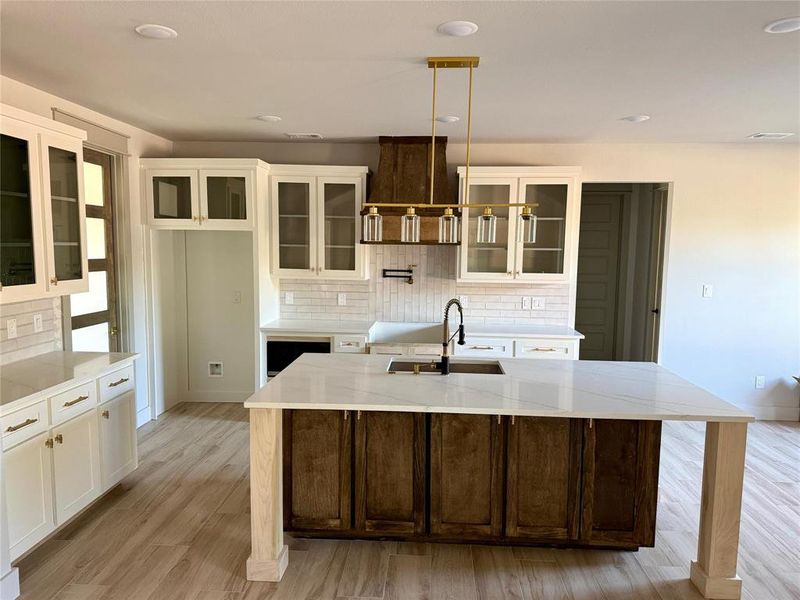 Kitchen featuring glass insert cabinets, white cabinetry, an island with sink, light stone counters, and backsplash