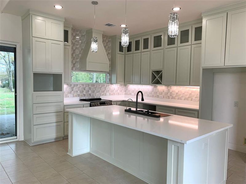 Kitchen featuring light tile patterned floors, custom range hood, a kitchen island with sink, tasteful backsplash, and decorative light fixtures Kitchen featuring light tile patterned floors, custom range hood, a kitchen island with sink, tasteful backsplash, and decorative light fixtures