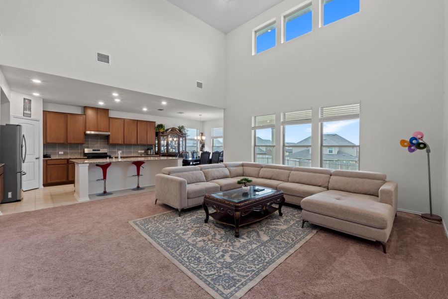 Open-concept living area with a two-story ceiling, abundant natural light, and a kitchen featuring wood-finish cabinetry, stainless steel appliances, and a tiled backsplash