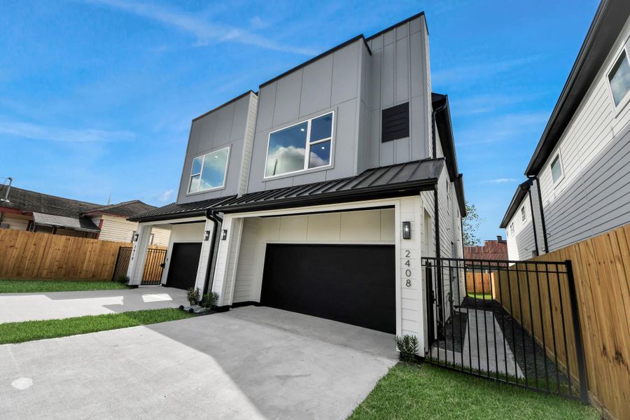This modern two-story home features a sleek exterior with large windows and a black metal roof. It includes a double garage, a gated pathway, and a fenced yard, offering both style and privacy.