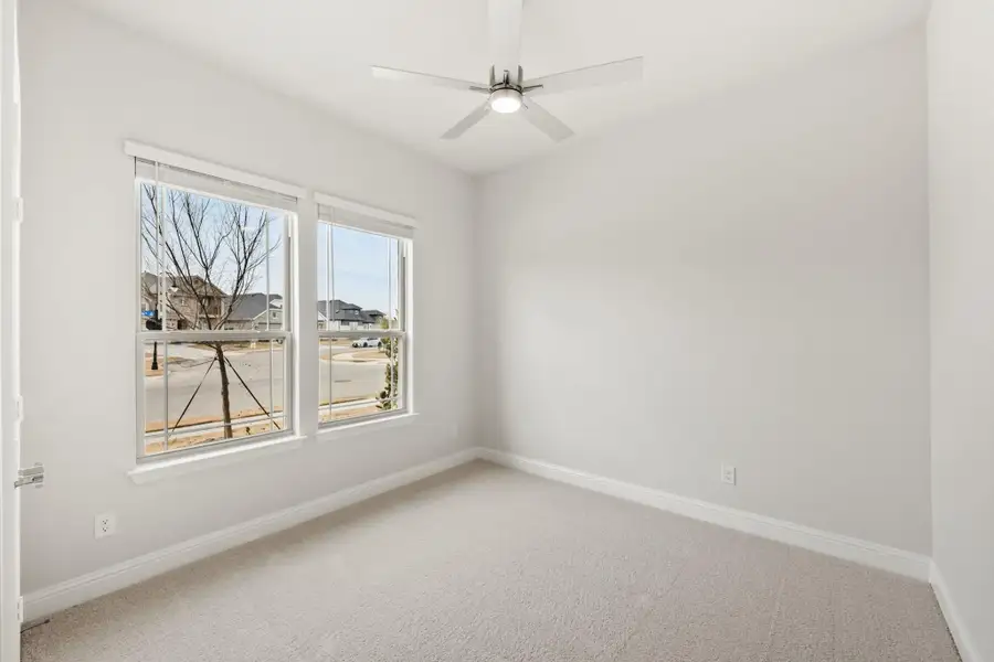 Empty bedroom with carpet flooring, ceiling fan, and two windows overlooking a neighborhood street Empty bedroom with carpet flooring, ceiling fan, and two windows overlooking a neighborhood street