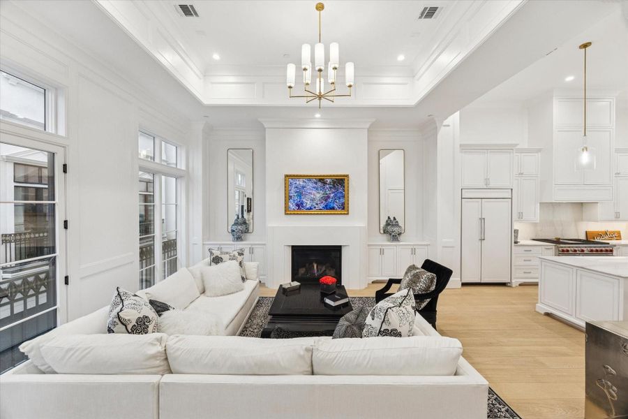 This is a spacious, elegant living room with 14' ceilings and a modern chandelier. It features a plaster finished fireplace, and an open layout connecting to a sleek kitchen.