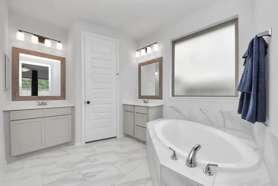 This bathroom features a modern design with a large soaking tub, dual vanities with framed mirrors, and elegant tile flooring. Frosted windows provide natural light while maintaining privacy.