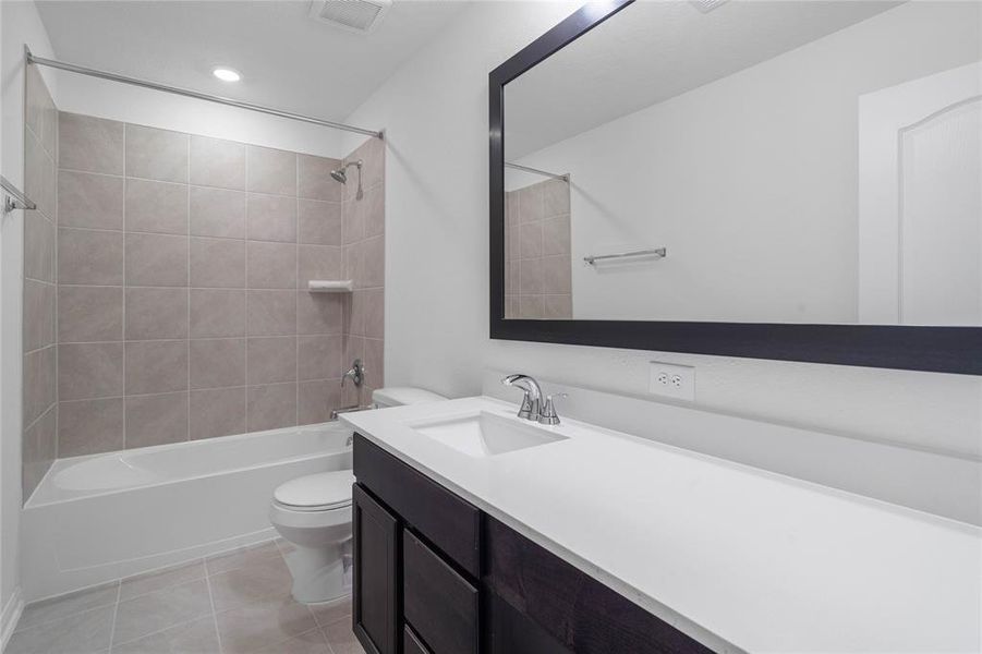 Secondary bath features tile flooring, bath/shower combo with tile surround, stained wood cabinets, beautiful light countertops, wood framed mirror, dark, sleek fixtures and modern finishes.