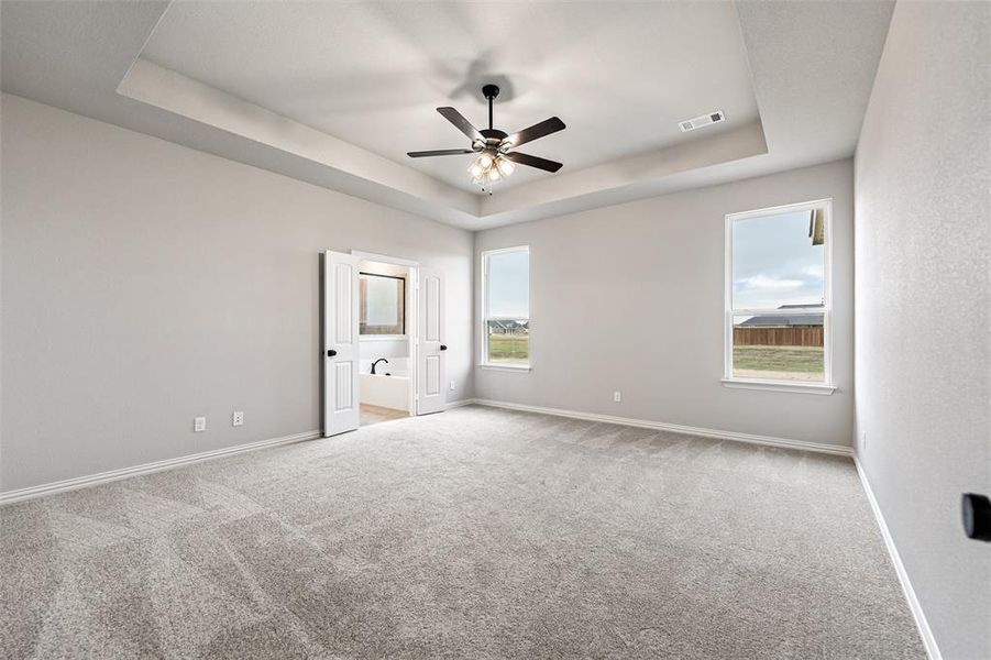 Unfurnished bedroom featuring a raised ceiling, light colored carpet, a ceiling fan, and ensuite bath Unfurnished bedroom featuring a raised ceiling, light colored carpet, a ceiling fan, and ensuite bath