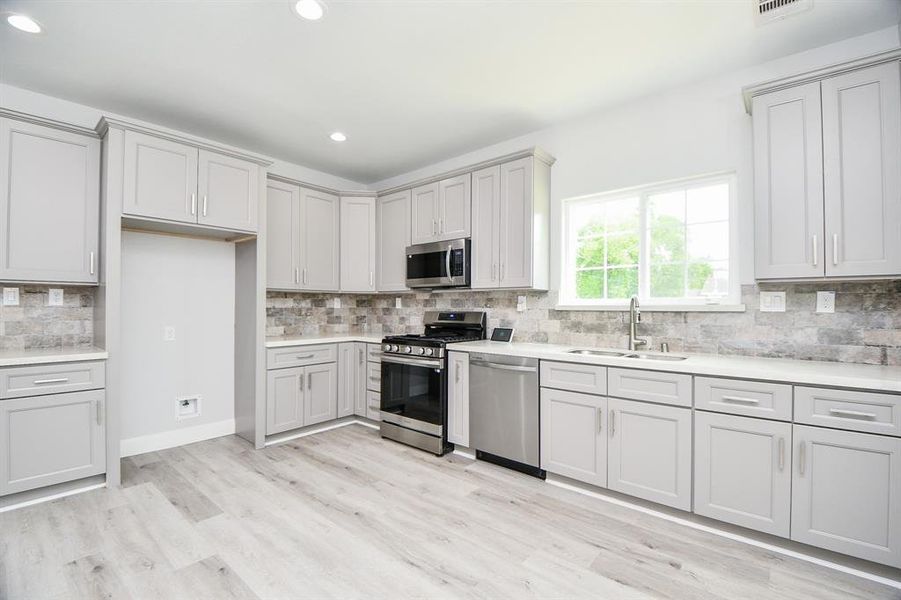 This is a modern, spacious kitchen featuring light gray cabinetry, stainless steel appliances, a stone backsplash, and light wood flooring, complemented by ample natural light from a large window.