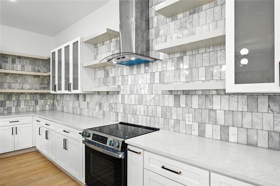 Kitchen with open shelves, stainless steel electric range oven, wall chimney exhaust hood, light countertops, and white cabinets Kitchen with open shelves, stainless steel electric range oven, wall chimney exhaust hood, light countertops, and white cabinets