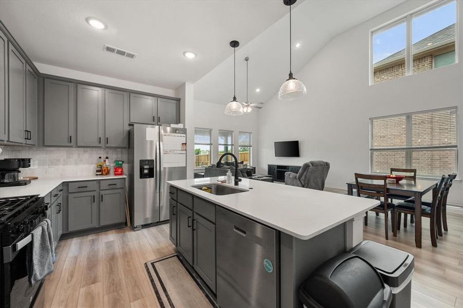 Open-concept kitchen featuring gray cabinetry, stainless steel appliances, a center island with sink, quartz countertops, and wood-finish flooring