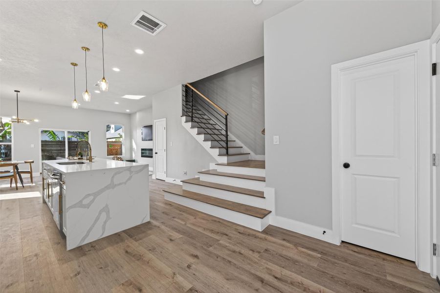 Chef’s kitchen open-concept living space anchored by a dramatic quartz waterfall island with bold veining, complemented by wide-plank white oak–finish laminate flooring throughout. A modern floating staircase with wood treads and sleek metal railing, designer pendant lighting, and recessed LED lights create a striking architectural focal point ideal for elevated entertaining.