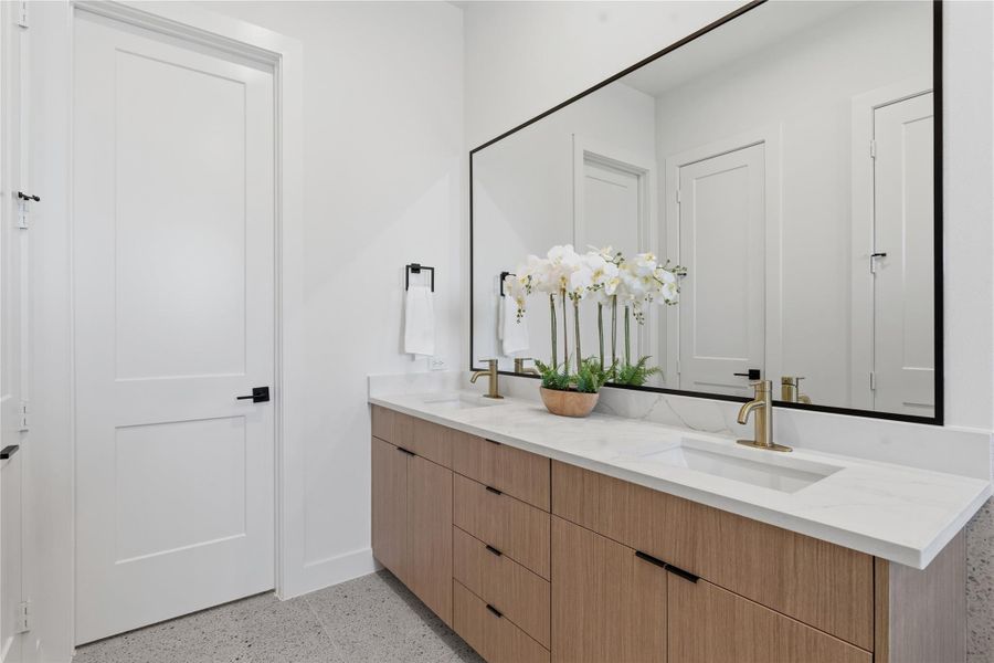 A double vanity with a light wood finish and white stone countertop anchors the bathroom. A large, black-framed mirror and brushed gold faucets provide sophisticated architectural accents.