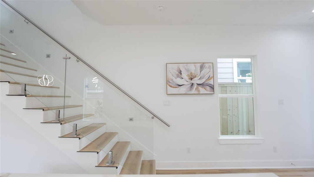 This photo showcases a modern interior with a sleek staircase featuring glass railings and light wood steps. The space is bright and airy, complemented by a large window and contemporary floral artwork on the wall, creating a clean and inviting atmosphere. This photo showcases a modern interior with a sleek staircase featuring glass railings and light wood steps. The space is bright and airy, complemented by a large window and contemporary floral artwork on the wall, creating a clean and inviting atmosphere.