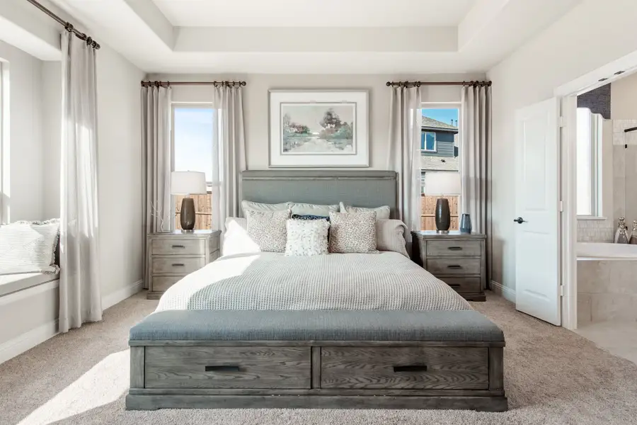 Master bedroom with king bed, gray storage frame, matching nightstands, tray ceiling, and ensuite bathroom door visible Master bedroom with king bed, gray storage frame, matching nightstands, tray ceiling, and ensuite bathroom door visible